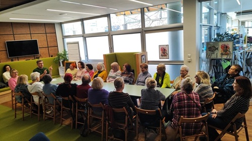 Grupa seniorek i senior&oacute;w wok&oacute;ł stołu konferencyjnego słucha z uwagą prowadzącego zajęcia