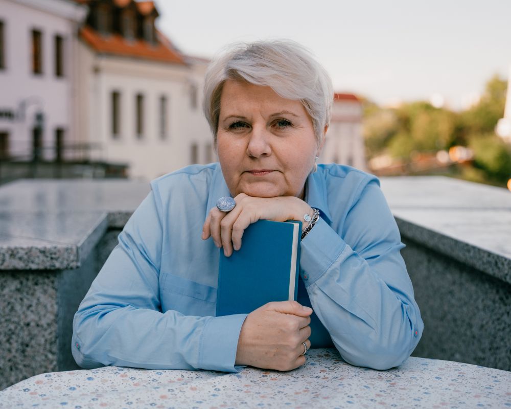 Alicja Filipowska - kobieta w średnim wieku, w krótkich, siwych włosach. Siedzi przy stole, trzyma w rękach książkę. 