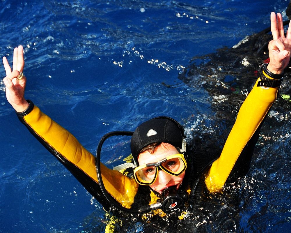 Justyna Szumacher - młoda kobieta w kostiumie płetwonurka będąca od szyi w dół w wodzie. Wynosi ręce w górę i palcami pokazuje znak "OK". 