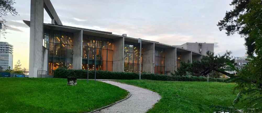 Zdjęcie przedstawia gmach biblioteki w Novej Goricy, w Słowenii. Przed budynkiem widać równoskoszoną trawę i prowadzącą do biblioteki drogę. 