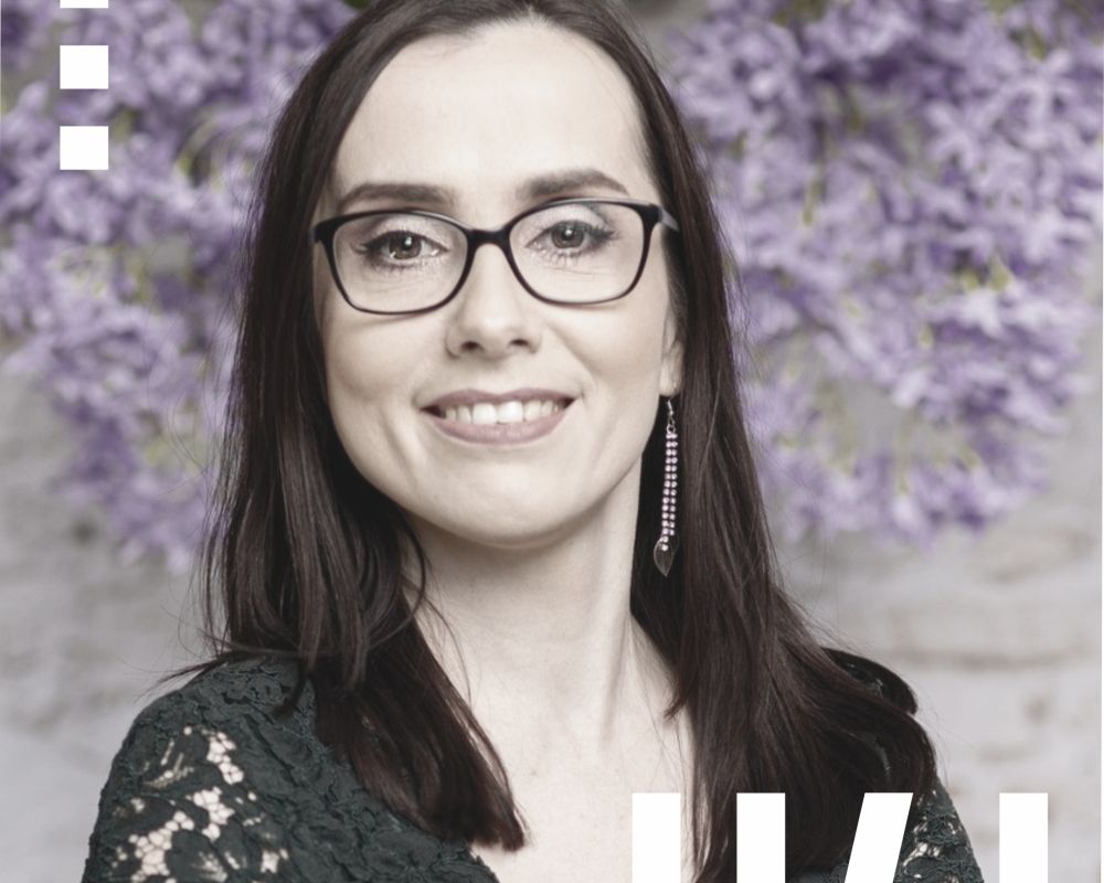 Anna Ziobro - młoda kobieta, w dłuższych, ciemnych włosach. Ma ubrane okulary z ciemnymi oprawkami. Siedzi na obróconym, białym krześle.