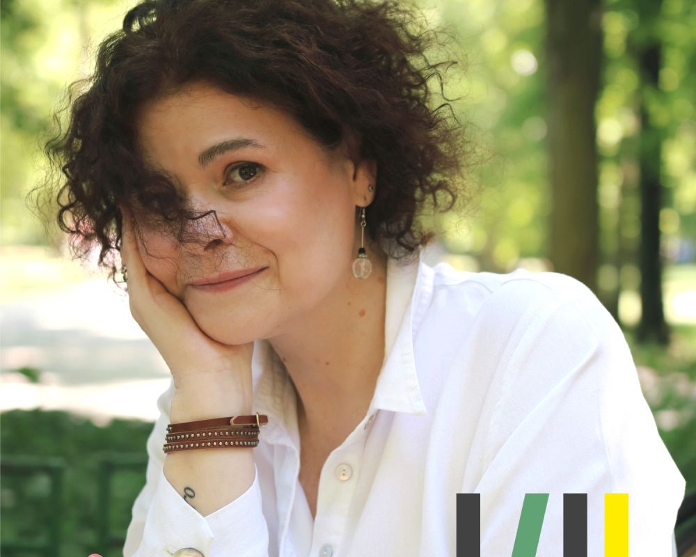 Anna Sakowicz - kobieta w kręconych włosach średniej długości. Siedząc, podpiera prawą ręką głowę. Ma na sobie białą koszulę. Na prawej ręce ma brązową bransoletkę.