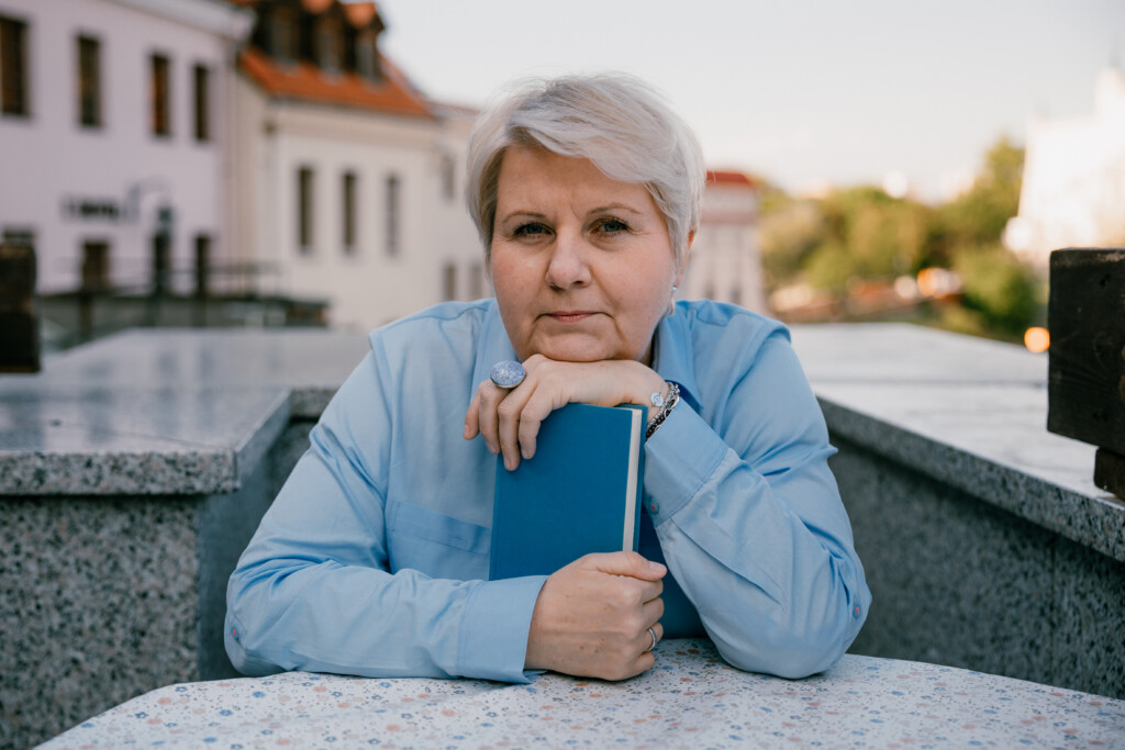 Alicja Filipowska - kobieta w średnim wieku, w krótkich, siwych włosach. Siedzi przy stole, trzyma w rękach książkę.
