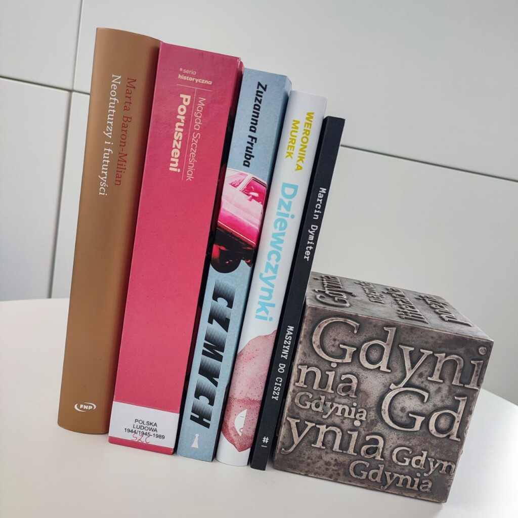 Zdjęcie przedstawia pięć stojących książek opartych o kostkę-nagrodę Nagrody Literackiej Gdynia, której widoczną ściankę pokrywają artystycznie rozmieszczone i różnej wielkości słowa "Gdynia".