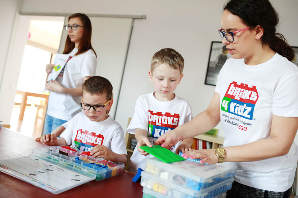 Dwie kobiety i dwóch chłopców układający klocki. Mają na sobie białe koszulki z logiem Bricks4Kids