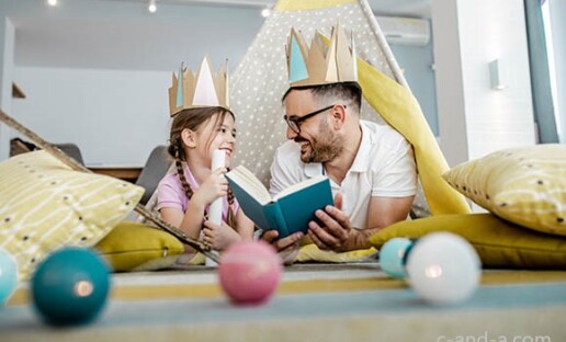 Dziewczynka i jej tata leżą na brzuchu na podłodze, podpierając się łokciami i wspólnie czytają książkę. Mają na głowach korony zrobione z papieru. Wokół widać rozrzucone poduszki i zabawki.