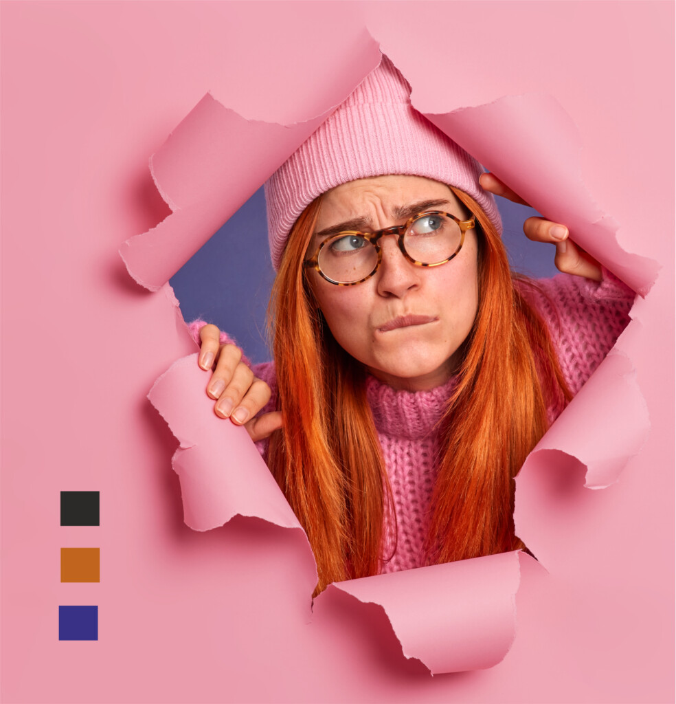Młoda kobieta w długich, rudych włosach. Ma na głowie różową czapkę, a na nosie okrągłe okulary. Kobieta patrzy w bok podejrzliwym wzrokiem. Wyłania się z dziury w różowej ścianie.