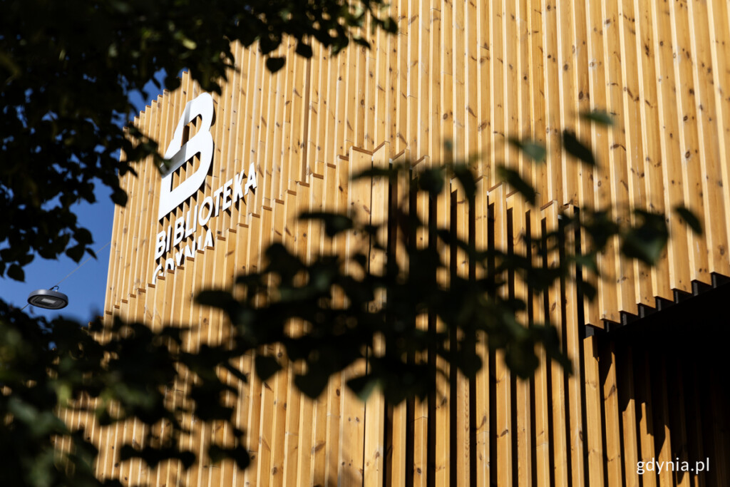 Biblioteka Witomino z zewnątrz. Elewacja z drewnianymi lamelami. W rogu budynku białe logo Biblioteki Gdynia
