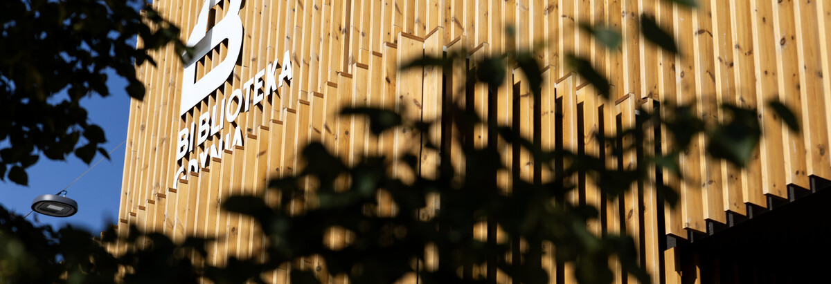 Zdjęcie przedstawia fasadę Biblioteki Witomino, na pierwszym planie znajdują się ciemne liście drzew.