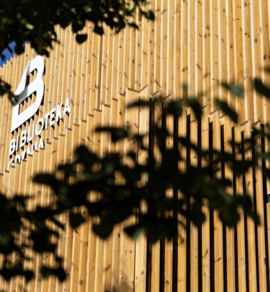 Zdjęcie przedstawia fasadę Biblioteki Witomino, na pierwszym planie znajdują się ciemne liście drzew.