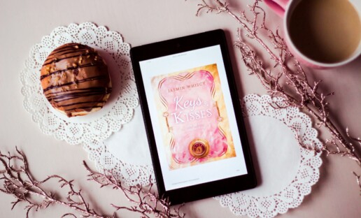 Włączony czytnik ebooków, na którym wyświetla się okładka książki. Leży na blacie, na którym leżą białe serwetki, kawa i ciastko czekoladowe.