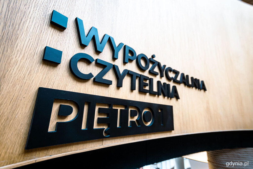 Czarny napis na drewnianej ściance "Wypożyczalnia. Czytelnia. Piętro 1"