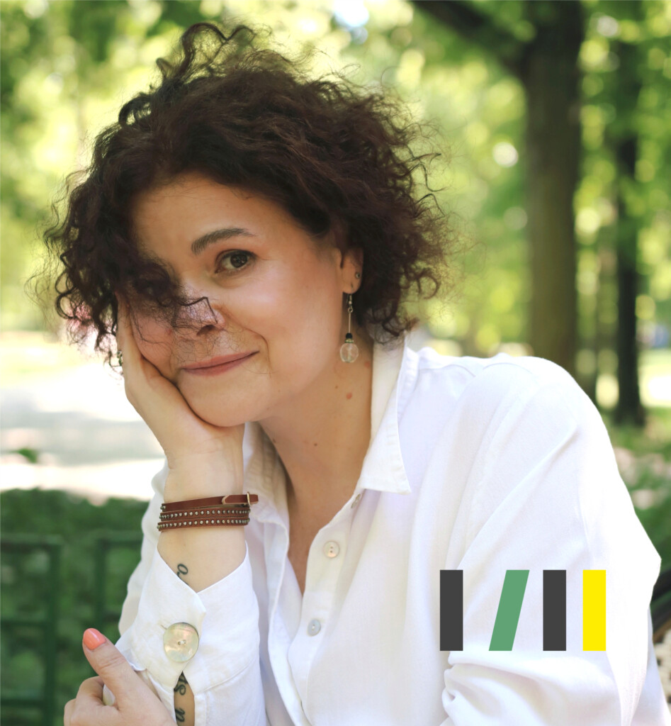 Anna Sakowicz - kobieta w kręconych włosach średniej długości. Siedząc, podpiera prawą ręką głowę. Ma na sobie białą koszulę. Na prawej ręce ma brązową bransoletkę.