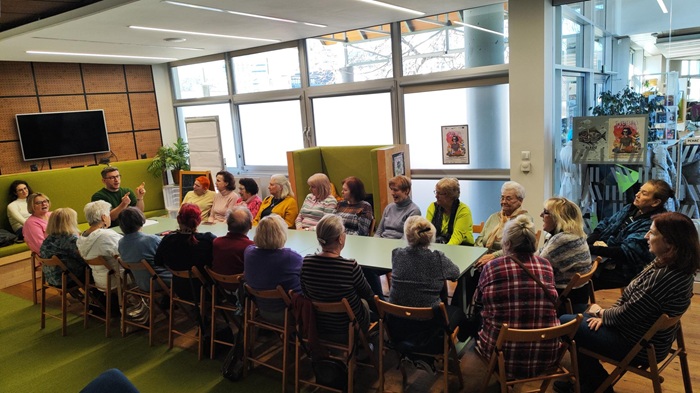 Grupa ok. 20 seniorek siedzi wok&oacute;ł stołu konferencyjnego i słucha z uwagą prowadzącego spotkanie.